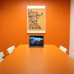 A conference room with an orange wall and an orange table to match it. There's an open laptop at the end of the conference table.