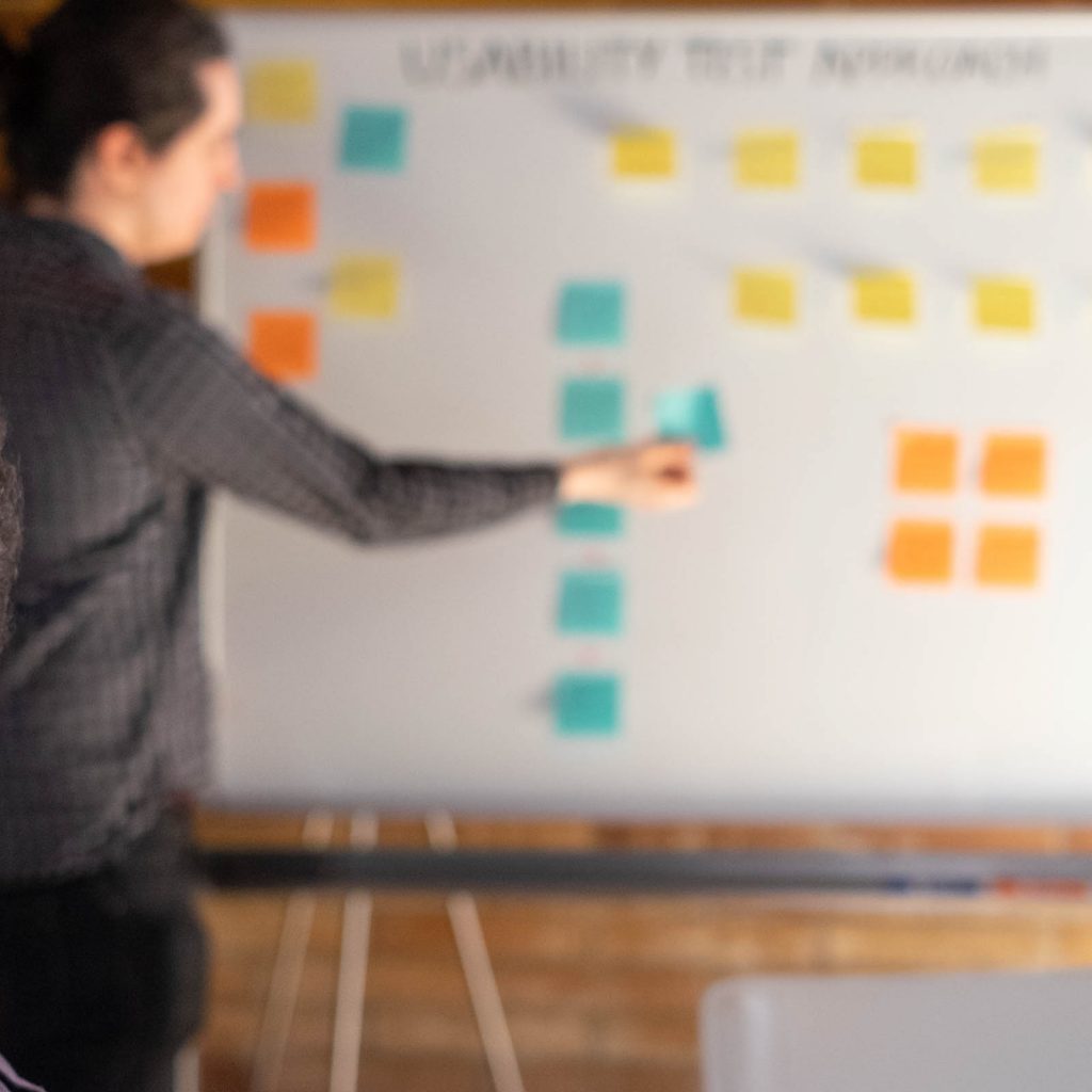 A person holding a post-it note up to a whiteboard, deciding where to start testing during the development life cycle.