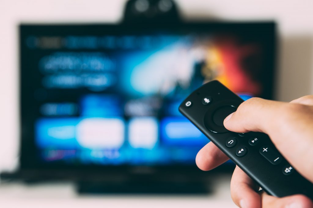 A hand holding the remote to streaming hardware, with the TV and streaming service info blurred behind it.