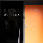 The front door of PLUS QA's office. It features a orange colored door with the number "1725" at the top. To the left of it is a tall black window with the PLUS QA logo painted on it.