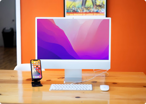 iMac device placed on a wood table. Next to it is an iPhone that is posed on a phone stand.