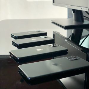A serious of iPhone 11 devices laying face down on a black, glossy desk. The computer monitor placed next to them can be seen in the reflection of the surface of the desk.