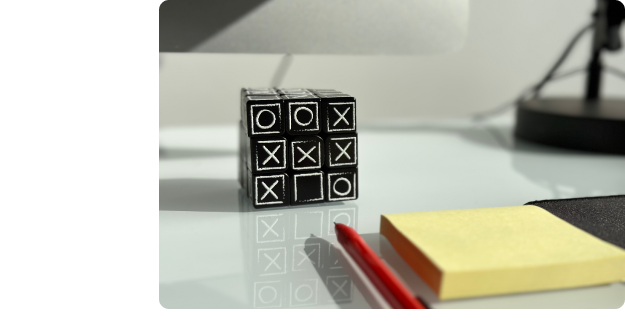 Close up view of a computer desk. In view is a 3x3 Rubiks cube that is painted black with white O and X symbols painted on each of the cube faces. There is also a sticky note pad with a pen.