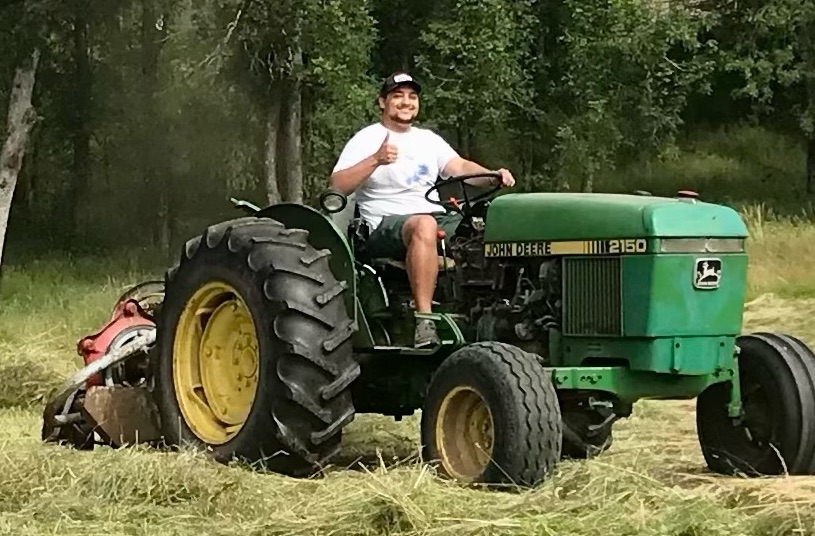 Brandon, QA analyst, smiling and giving a thumbs up while driving a large riding tractor on his farm.
