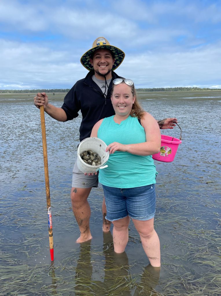 Brandon and his fiancee on a beach, smiling and holding up buckets full of clams.