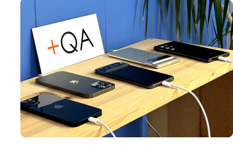 A collection of mobile devices arranged on a shelf. Placed next to it is a sign that reads "+QA"